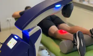Pacjent leży na kozetce, a na jego kolanie działa nowoczesny laser wysokoenergetyczny, emitując czerwone światło.