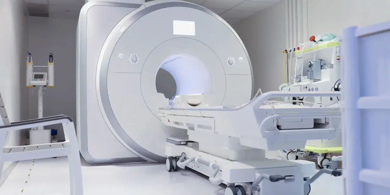 Nowoczesny gabinet diagnostyczny z aparaturą do rezonansu kręgosłupa szyjnego. Obok leżanka i sprzęt medyczny.
