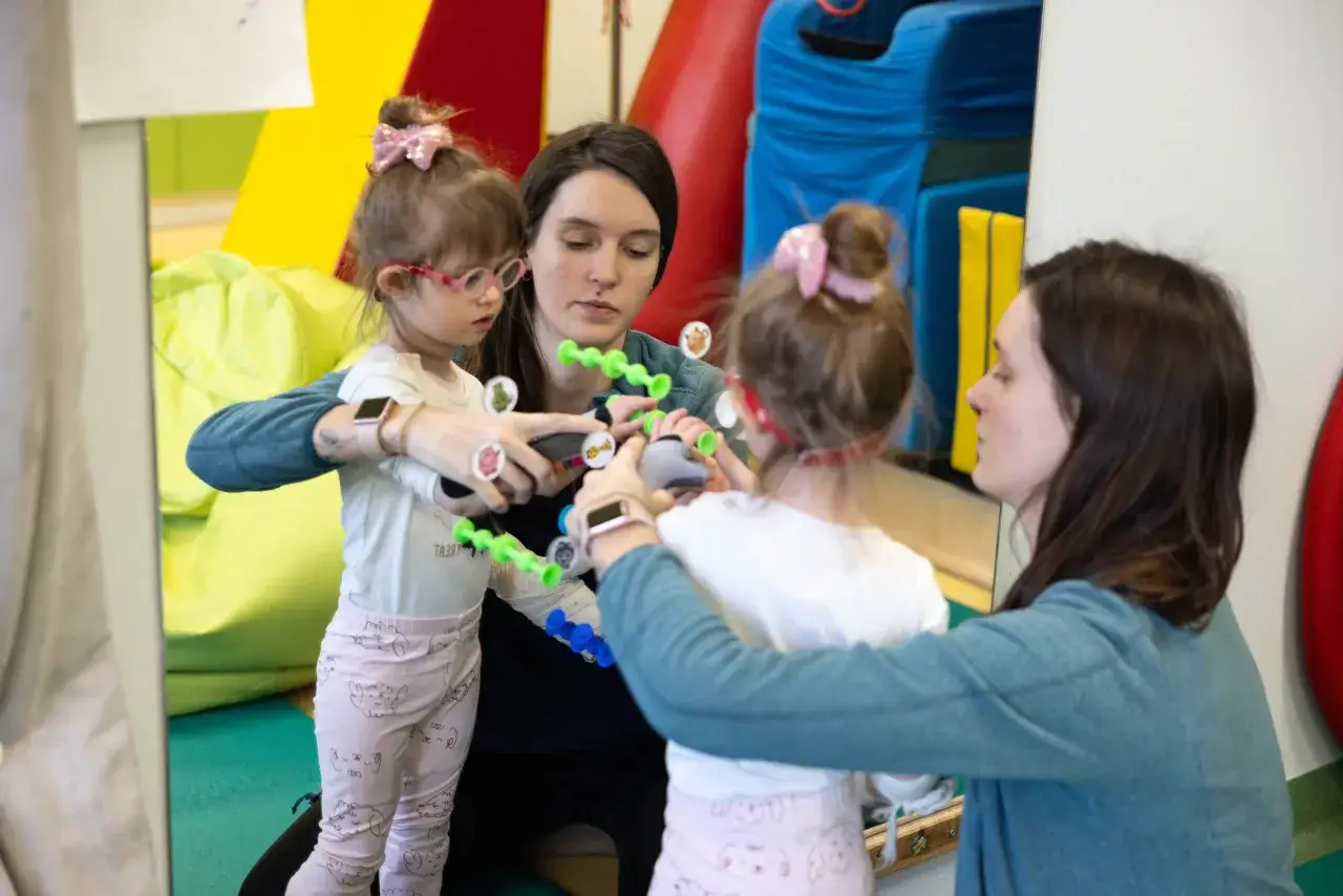 Dwie kobiety pomagają dziewczynce w okularach w zabawie koralikami. To przykład holistyczne podejście do rozwoju dziecka.