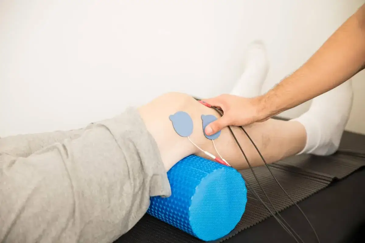 Rehabilitacja po artroskopii kolana: elektrody EMS na udzie, wałek do masażu pod kolanem, fizjoterapeuta.
