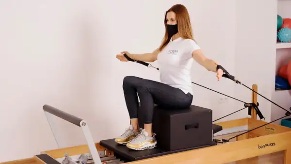 Kobieta w masce ćwiczy na reformerze pilates, wykonując ćwiczenie z linkami.